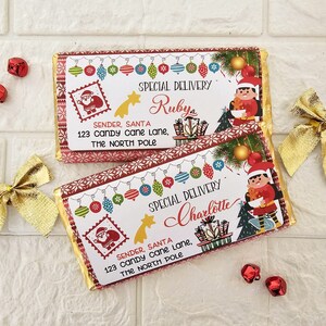 Może przedstawiać: Dwie czerwono-białe czekoladowe tabliczki z motywem świątecznym i świątecznym wzorem. Górna tabliczka zawiera napis "Special Delivery Ruby", a dolna "Special Delivery Charlotte". Na obu tabliczkach widnieje Święty Mikołaj i elf.