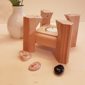 Könnte beinhalten: Ein Kerzenhalter aus Holz mit vier Beinen und einer quadratischen Basis. Eine kleine weiße Kerze sitzt in der Mitte der Basis. Der Halter ist aus hellem Holz gefertigt und hat eine natürliche Oberfläche.
