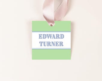 May include: A light green gift tag with the name "EDWARD TURNER" in blue text. The text is centered on a white rectangular band with blue borders. A pink ribbon is attached to the top of the tag.