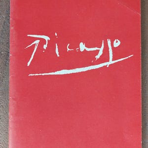 Op de afbeelding: Een rechthoekig boek met een effen rode kaft met het woord Picasso in een wit, gestileerd, handgeschreven lettertype in het midden. Het boek heeft een verticale rug aan de linkerkant en een strakke, matte afwerking op de voorzijde van de kaft.