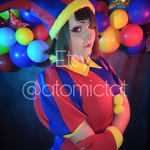 Puede incluir: Una persona que lleva un traje de bufón rojo, azul y amarillo con un sombrero puntiagudo. La persona está de pie frente a una pared decorada con globos de colores.