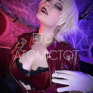 Peut inclure: Une personne porte un costume de cosplay avec une veste rouge, une bordure en dentelle noire et des gants blancs. La personne a les cheveux blonds et est maquillée. L'arrière-plan a une palette de couleurs violet et rose avec des toiles d'araignées. Le texte "etsy atomictot" est visible.