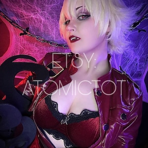 May include: A person in a cosplay outfit with a red jacket, a red and black top, and a blonde wig. The image has the text "ETSY" and "ATOMICTOT". The background is decorated with cobwebs and chains.
