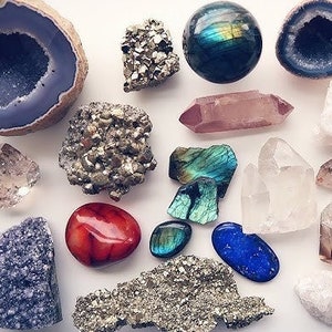 Puede incluir: Una colección de varios cristales y piedras, incluyendo amatista, cuarzo rosa, labradorita, cuarzo transparente, pirita y ágata. Los cristales están dispuestos sobre una superficie blanca.