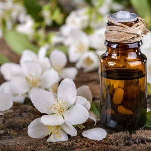 May include: A small amber glass bottle with a cork stopper and a brown twine tied around the neck. The bottle is filled with a clear liquid and is surrounded by white jasmine flowers on a wooden surface.
