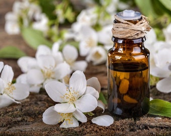 May include: A small amber glass bottle with a cork stopper and a brown twine tied around the neck. The bottle is filled with a clear liquid and is surrounded by white jasmine flowers on a wooden surface.