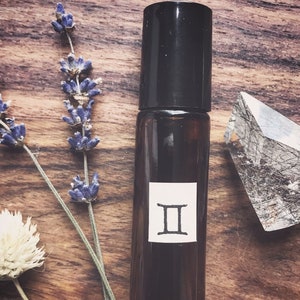May include: A brown glass roller bottle with a black cap and a white label featuring the Gemini zodiac symbol. The bottle is on a wooden surface with lavender flowers and a clear crystal.