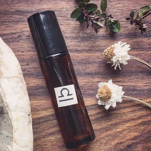 May include: A brown glass roller bottle with a black cap and a white label featuring the Libra zodiac symbol. The bottle is on a wooden surface with dried flowers.