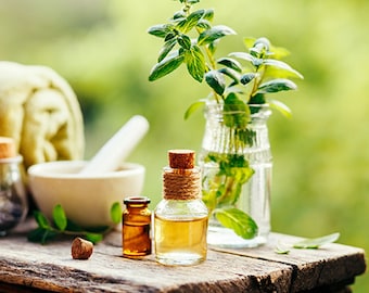 May include: Four small glass bottles with cork tops filled with essential oils, a white ceramic mortar and pestle, and a glass vase with fresh green herbs. The bottles and vase are sitting on a rustic wooden surface.