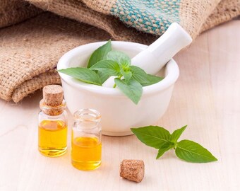 May include: Two small glass bottles with cork tops filled with a yellow liquid, a white mortar and pestle with green basil leaves inside, and a sprig of basil leaves on a light wood surface.