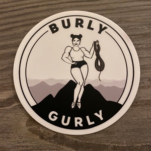 Puede incluir: Una pegatina redonda blanca con un contorno negro. La pegatina presenta una ilustración en blanco y negro de una mujer de pie en una montaña con una cuerda en la mano. El texto "Burly Gurly" está escrito en letras negras alrededor del borde de la pegatina.