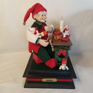 May include: A Christmas elf figurine wearing a red hat and coat, green pants, and white collar. The elf is holding a toy and sitting on a red box. A small table holds a candle, rocking horse, and wreath. The base reads "NOEL".