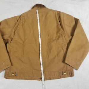 Puede incluir: Una chaqueta de trabajo marr&oacute;n con cuello marr&oacute;n, vista desde atr&aacute;s. Una cinta m&eacute;trica se coloca verticalmente en el centro de la chaqueta, indicando su longitud. La chaqueta tiene dos bolsillos delanteros y botones met&aacute;licos. La chaqueta mide unos 63.5 cm de largo.