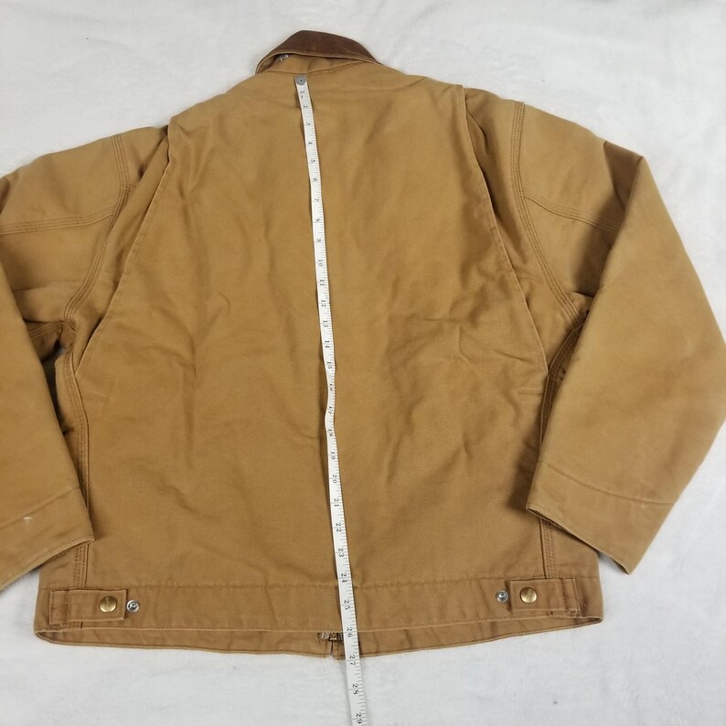 Puede incluir: Una chaqueta de trabajo marr&oacute;n con cuello marr&oacute;n, vista desde atr&aacute;s. Una cinta m&eacute;trica se coloca verticalmente en el centro de la chaqueta, indicando su longitud. La chaqueta tiene dos bolsillos delanteros y botones met&aacute;licos. La chaqueta mide unos 63.5 cm de largo.