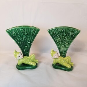 May include: A pair of green ceramic vases with a whimsical design. Each vase features a reclining elf figure with a green and white painted design. The vases have a fan-shaped opening and are decorated with green leaves and flowers.