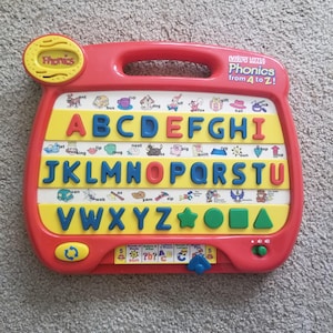 May include: A red VTech Little Smart Phonics from A to Z learning toy featuring a yellow panel with the alphabet in blue and red raised letters, small illustrations for each letter, and a bottom row with green geometric shapes and control buttons.