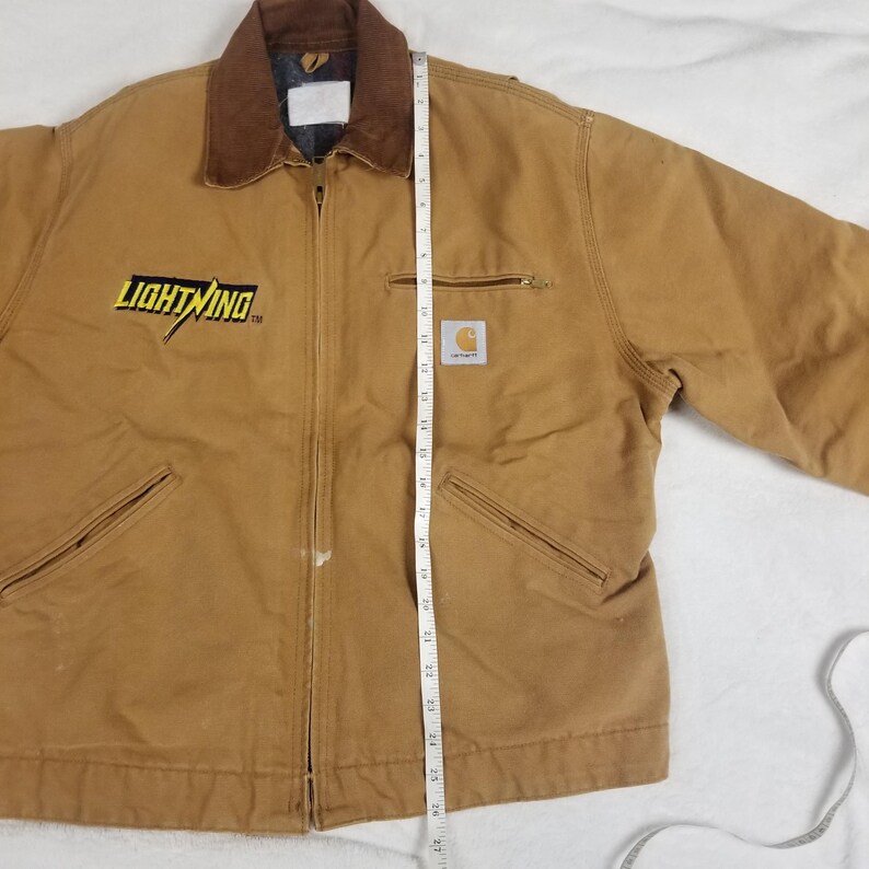 Puede incluir: Chaqueta de trabajo Carhartt de color marr&oacute;n con cuello de pana marr&oacute;n. La chaqueta tiene cierre de cremallera, dos bolsillos delanteros y la palabra "LIGHTNING" en letras amarillas. La chaqueta mide unos 66 cm de largo.