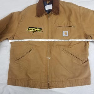 Puede incluir: Chaqueta de trabajo Carhartt de color marr&oacute;n con cuello de pana marr&oacute;n. La chaqueta tiene cierre de cremallera, dos bolsillos delanteros y un bolsillo en el pecho. La palabra "LIGHTNING" est&aacute; en letras amarillas en el pecho izquierdo. El logotipo de Carhartt est&aacute; en el pecho derecho.