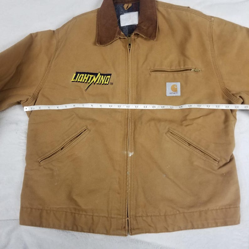 Puede incluir: Chaqueta de trabajo Carhartt de color marr&oacute;n con cuello de pana marr&oacute;n. La chaqueta tiene cierre de cremallera, dos bolsillos delanteros y un bolsillo en el pecho. La palabra "LIGHTNING" est&aacute; en letras amarillas en el pecho izquierdo. El logotipo de Carhartt est&aacute; en el pecho derecho.