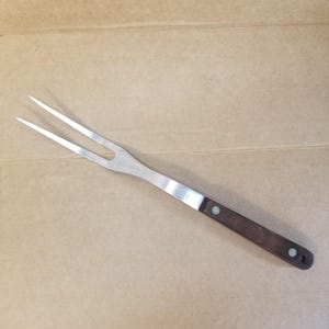 May include: A stainless steel carving fork with two tines. The handle is dark brown wood with two small, round, green-colored rivets. The fork is designed for handling and serving cooked meats.