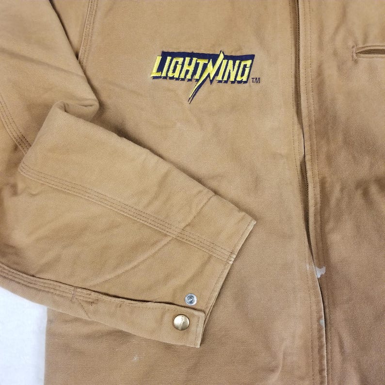 Puede incluir: Primer plano de una chaqueta de trabajo color beige con la palabra "LIGHTNING" bordada en amarillo y azul. La chaqueta tiene un pu&ntilde;o abotonado y un bolsillo. La tela parece duradera y adecuada para el trabajo.