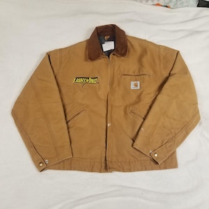 Puede incluir: Chaqueta de trabajo de color marr&oacute;n con cuello de pana marr&oacute;n. La chaqueta tiene la palabra "LIGHTNING" en letras amarillas y negras en el pecho izquierdo y un logotipo de Carhartt en el pecho derecho. Tiene dos bolsillos delanteros y una cremallera.