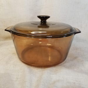 May include: A round, transparent amber-colored glass casserole dish with a matching glass lid. The dish features two integrated side handles and a central knob on the lid. The glass has a smooth, polished finish and a deep brown tint throughout.