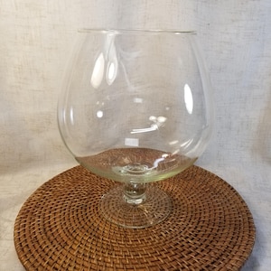 May include: A large, clear glass bowl with a wide, rounded body and a short, sturdy stem that connects to a circular base. The glass is transparent and smooth, featuring a wide opening at the top and a thick, solid foot for stability.
