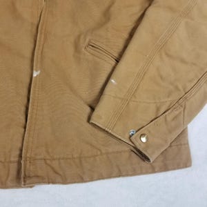 Puede incluir: Primer plano de una chaqueta de trabajo color canela. La chaqueta tiene un pu&ntilde;o abotonado, un peque&ntilde;o bolsillo y detalles de costura visibles. Hay algunas peque&ntilde;as marcas de pintura blanca en la chaqueta. La tela parece ser un material duradero y resistente.