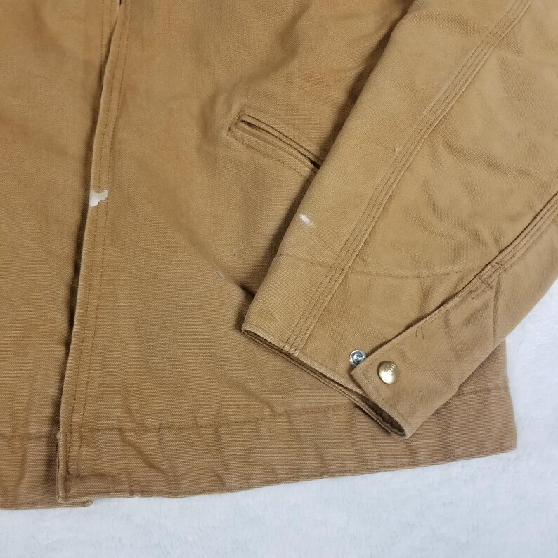 Puede incluir: Primer plano de una chaqueta de trabajo color canela. La chaqueta tiene un pu&ntilde;o abotonado, un peque&ntilde;o bolsillo y detalles de costura visibles. Hay algunas peque&ntilde;as marcas de pintura blanca en la chaqueta. La tela parece ser un material duradero y resistente.