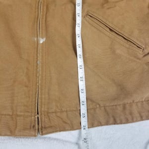 Puede incluir: Primer plano de una chaqueta de trabajo color canela con cierre de cremallera y un bolsillo. Una cinta m&eacute;trica se coloca verticalmente a lo largo de la chaqueta, mostrando medidas en cent&iacute;metros. La chaqueta tiene algunas manchas blancas.