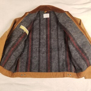 Puede incluir: Una chaqueta de trabajo marr&oacute;n con cuello de pana marr&oacute;n. El forro interior es gris con rayas verticales rojas y negras. La chaqueta tiene un peque&ntilde;o bolsillo con una etiqueta amarilla y una etiqueta blanca.