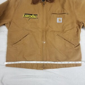 Puede incluir: Chaqueta de trabajo marr&oacute;n con cuello de pana marr&oacute;n. La chaqueta presenta el texto "LIGHTNING" en amarillo y un logotipo de Carhartt. Tiene cierre de cremallera, dos bolsillos delanteros y un bolsillo en el pecho.
