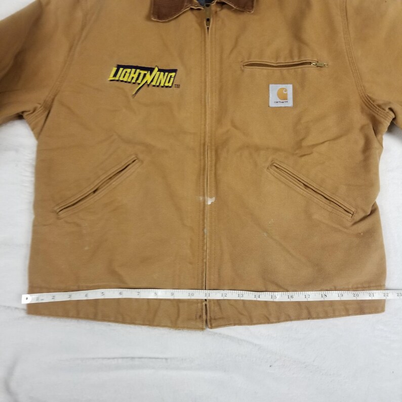 Puede incluir: Chaqueta de trabajo marr&oacute;n con cuello de pana marr&oacute;n. La chaqueta presenta el texto "LIGHTNING" en amarillo y un logotipo de Carhartt. Tiene cierre de cremallera, dos bolsillos delanteros y un bolsillo en el pecho.