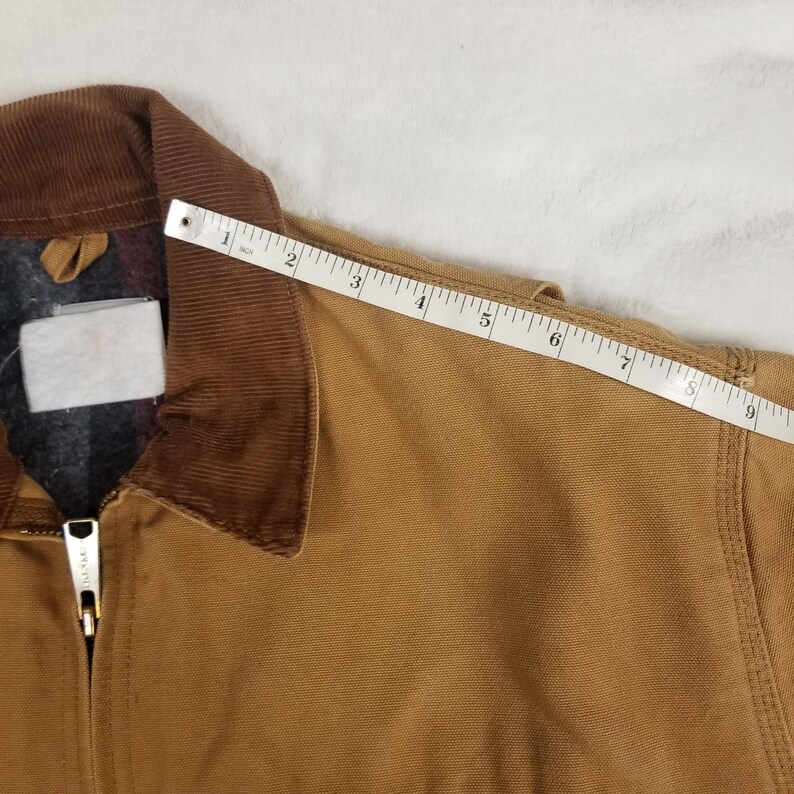 Puede incluir: Chaqueta de lona marr&oacute;n con cuello de pana y forro a cuadros. La chaqueta tiene cierre de cremallera y una etiqueta blanca. Una cinta m&eacute;trica muestra una longitud de aproximadamente 23 cm.