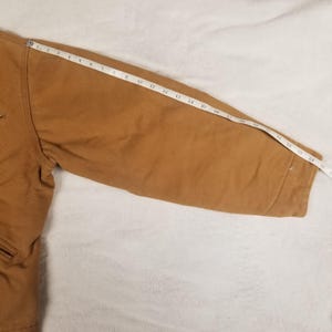 Puede incluir: Una manga de chaqueta de trabajo de color marr&oacute;n con una cinta m&eacute;trica colocada encima. La manga es de color marr&oacute;n liso. La cinta m&eacute;trica muestra una longitud de manga de aproximadamente 63,5 cm.