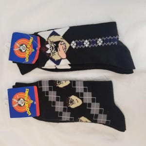 May include: Two pairs of black socks with a white and gray argyle pattern. One pair features a cartoon image of Tasmanian Devil from Looney Tunes. The other pair features a cartoon image of Tweety Bird from Looney Tunes. The socks are packaged with a blue and white Looney Tunes label.