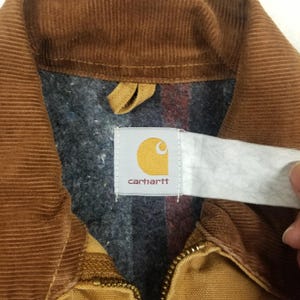 Puede incluir: Primer plano del cuello de una chaqueta de pana marr&oacute;n con forro a cuadros gris y rojo. La chaqueta tiene una cremallera y una etiqueta Carhartt con un logotipo amarillo y el nombre de la marca.