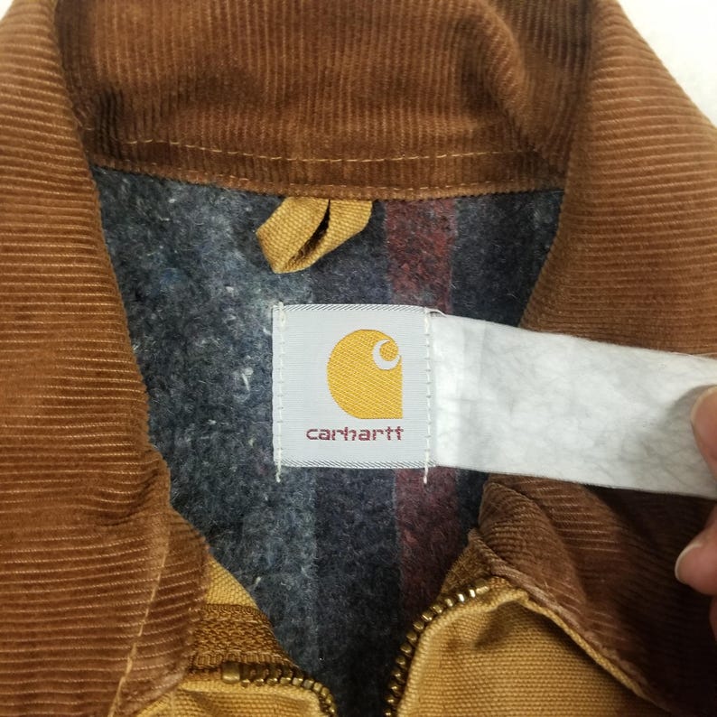 Puede incluir: Primer plano del cuello de una chaqueta de pana marr&oacute;n con forro a cuadros gris y rojo. La chaqueta tiene una cremallera y una etiqueta Carhartt con un logotipo amarillo y el nombre de la marca.
