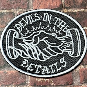 Op de afbeelding: Zwart-wit geborduurde patch met de tekst "Devils in the Details" in een cirkel. De patch heeft een handdrukdesign.