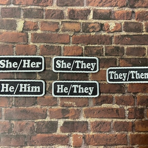 Peut inclure: Six patchs rectangulaires noirs avec du texte blanc. Les patchs indiquent "She/Her", "She/They", "They/Them", "He/Him", et "He/They".