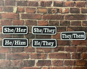 Può includere: Sei toppe rettangolari nere con testo bianco. Le toppe recitano "She/Her", "She/They", "They/Them", "He/Him", e "He/They".