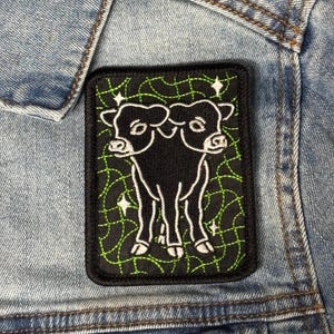 May include: A black, rectangular patch with a white outline of a two-headed cow. The background is black with a green, web-like pattern and white star accents. The patch is attached to a denim jacket.