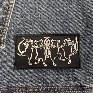 May include: A rectangular black embroidered patch featuring a white line art design of four rats standing in a circle and holding tails. The patch is securely attached to a blue denim fabric background with visible stitching along the edges.