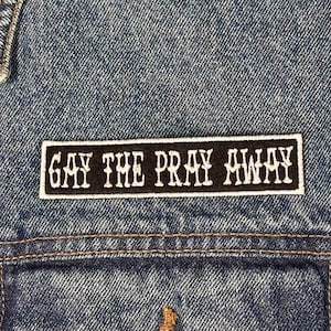 Peut inclure: Gros plan sur une veste en jean avec un écusson rectangulaire. L'écusson affiche le texte "GAY THE PRAY AWAY" en blanc sur fond noir. L'écusson est cousu sur le devant de la veste.