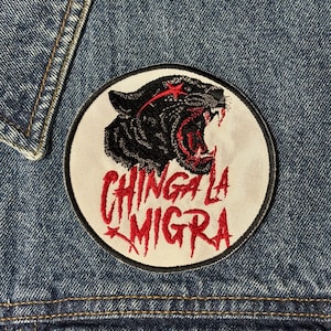 May include: Embroidered patch featuring a black panther with red accents and the text "CHINGA LA MIGRA" in red. The patch is circular with a white background and a black border, sewn onto a denim jacket.