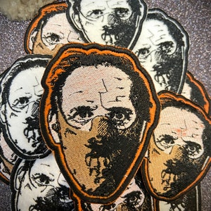 May include: A stack of embroidered patches featuring a stylized image of a person wearing a mask. The patches are black, white, and orange with a detailed design.
