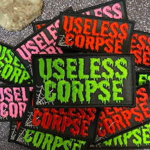 Op de afbeelding: Een stapel zwarte stoffen patches met de woorden "Useless Corpse" in groene, rode en roze letters. De patches hebben een spinnenwebontwerp in wit garen.