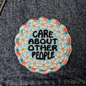 Può includere: Toppa ricamata con la scritta "CARE ABOUT OTHER PEOPLE" in nero su sfondo azzurro. La toppa ha un bordo floreale con margherite bianche e un bordo smerlato rosa, blu e crema. La toppa è su una giacca di jeans.