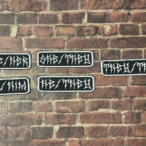 May include: Five black and white embroidered pronoun patches. Each patch has a white border and features text such as "SHE/HER", "SHE/THEY", "THEY/THEM", "HE/HIM", and "HE/THEY". The patches are displayed against a brick wall background.